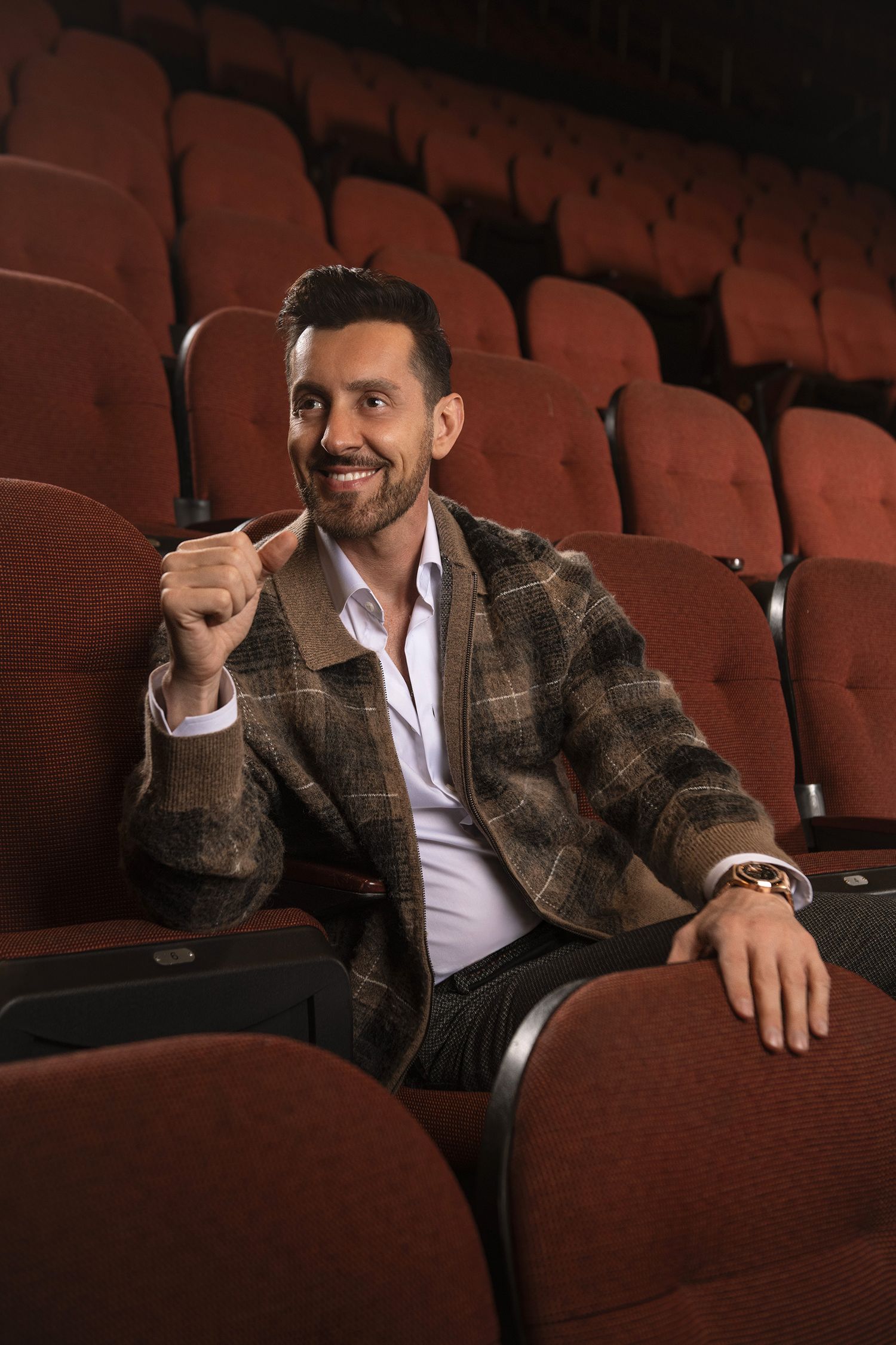 Entertainer and comedian Max Amini in plaid jacket sits relaxed in theater seat with confident smile in kia forum.