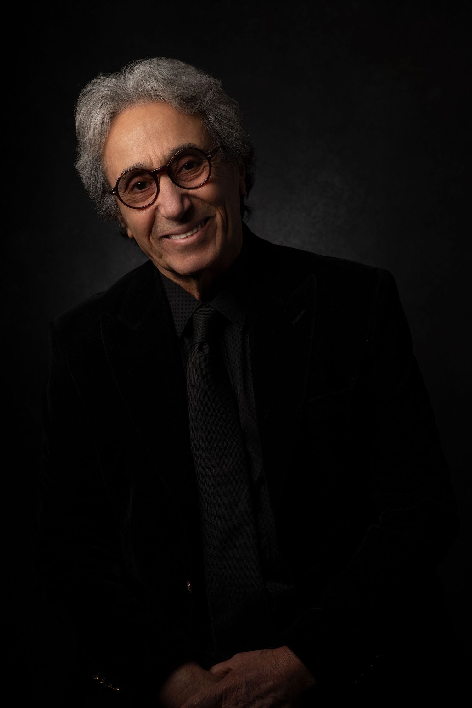 Portrait series of persian pop singer Shahbal Shabpareh with gray hair and glasses wearing dark clothing against black background.