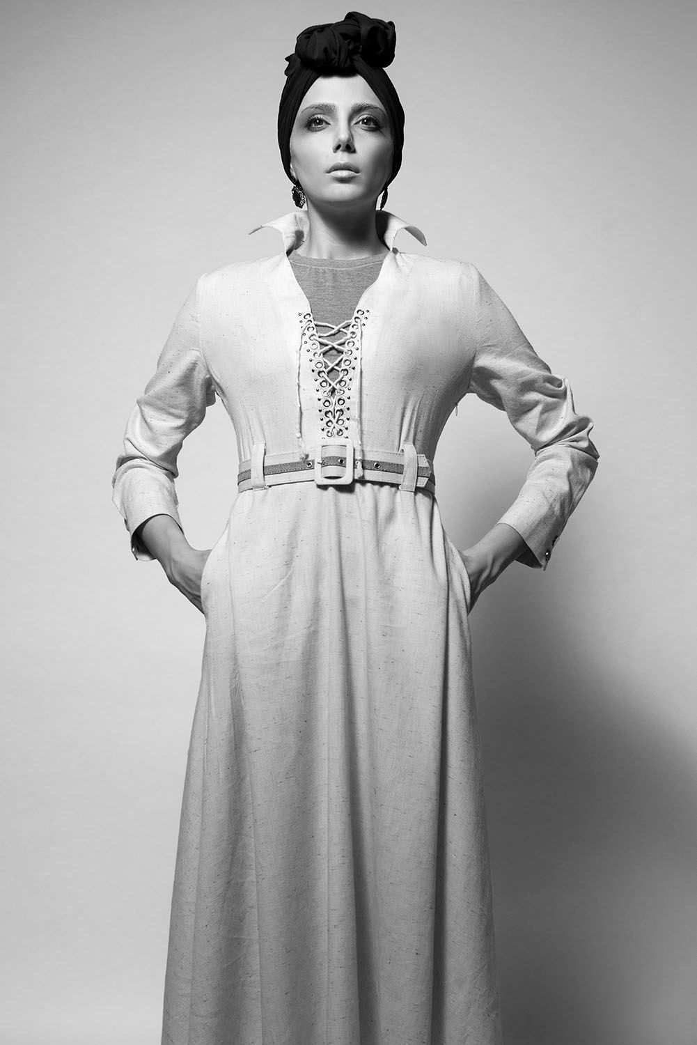 Black and white fashion photo of a woman in a long white belted dress with lace-up neckline and elegant updo hairstyle.