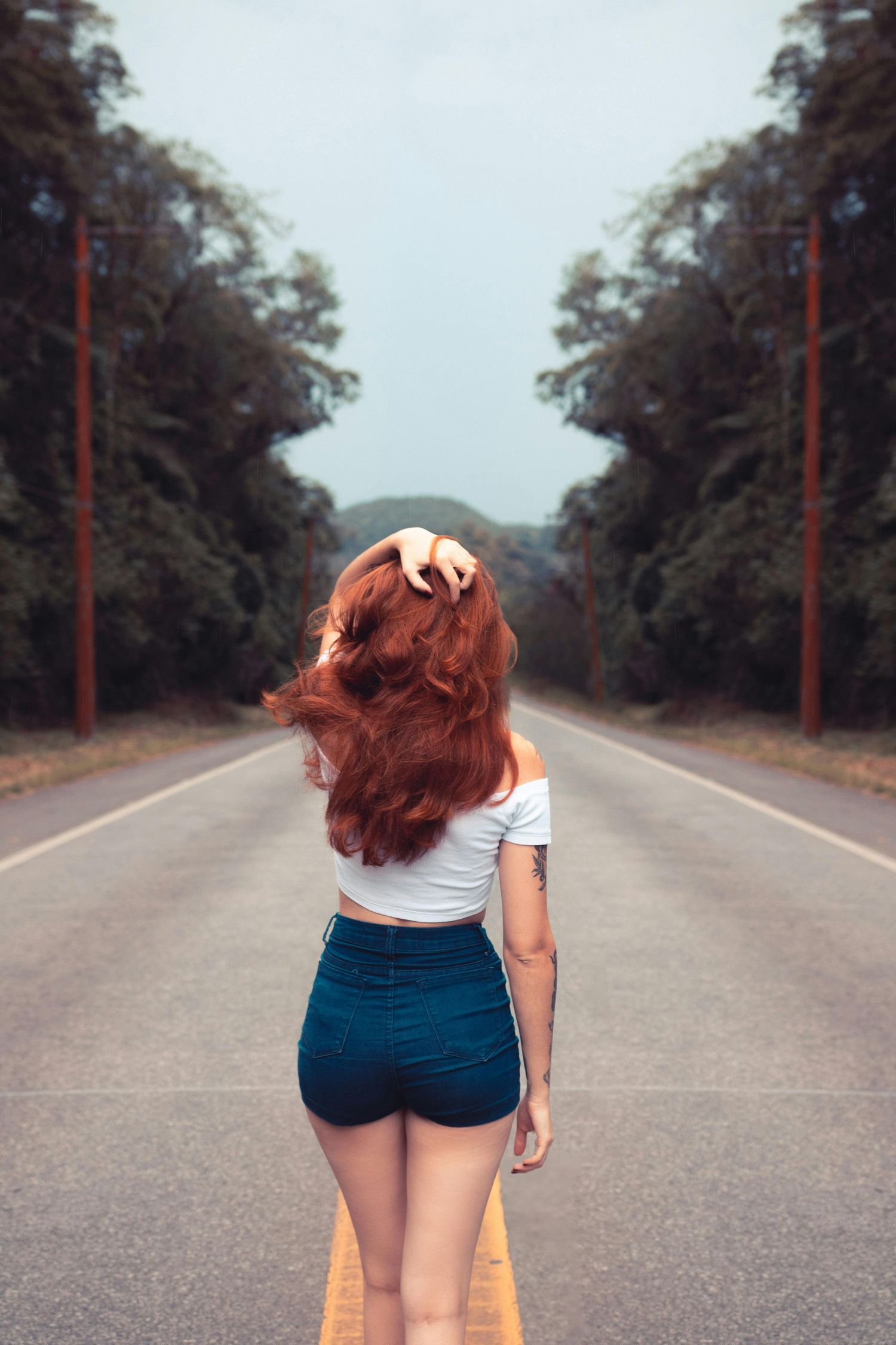 Person with red hair walking away on an empty tree-lined road.