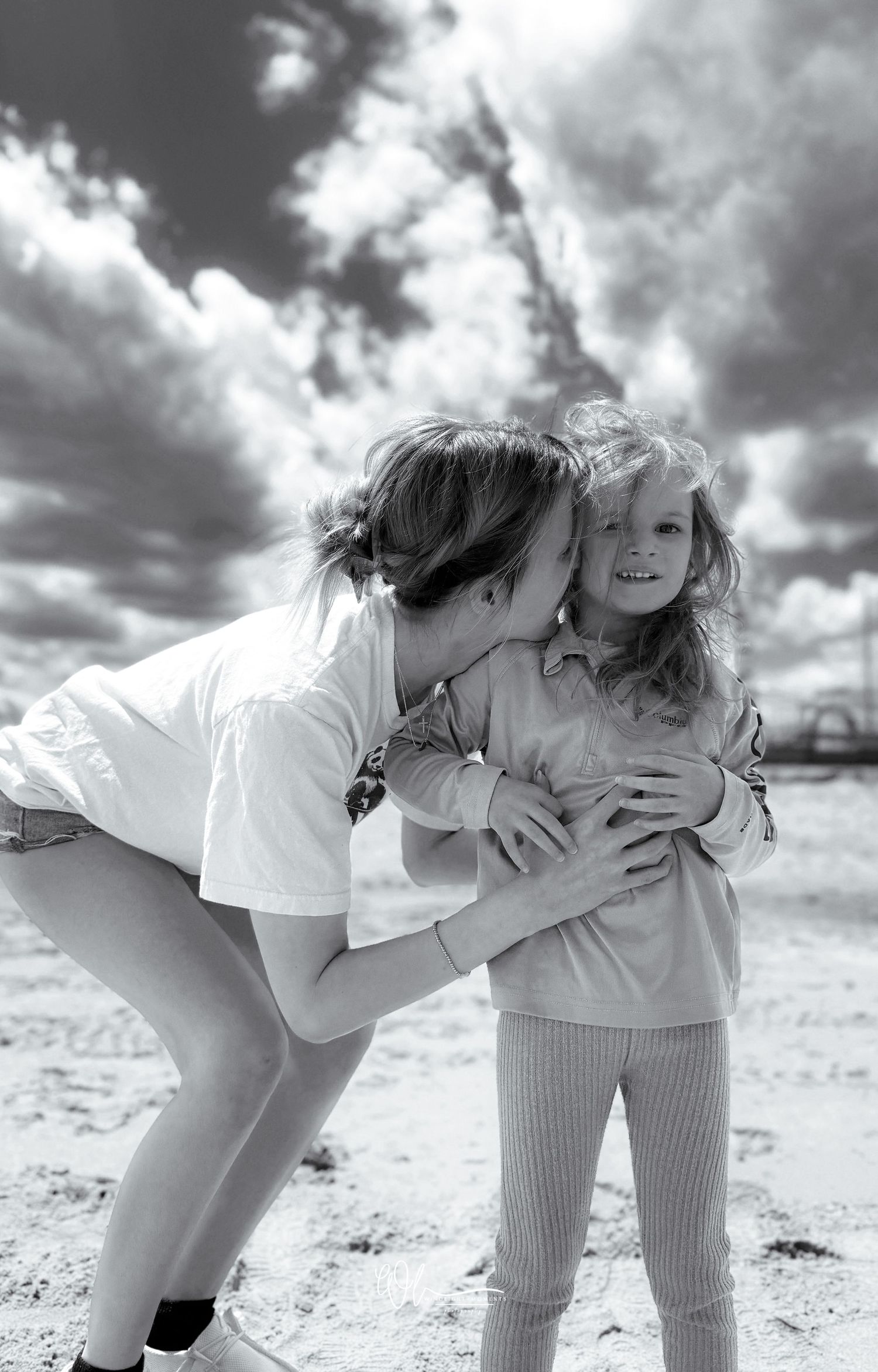 A heartwarming black and white moment of affection and laughter captured outdoors against dramatic cloudy skies.
