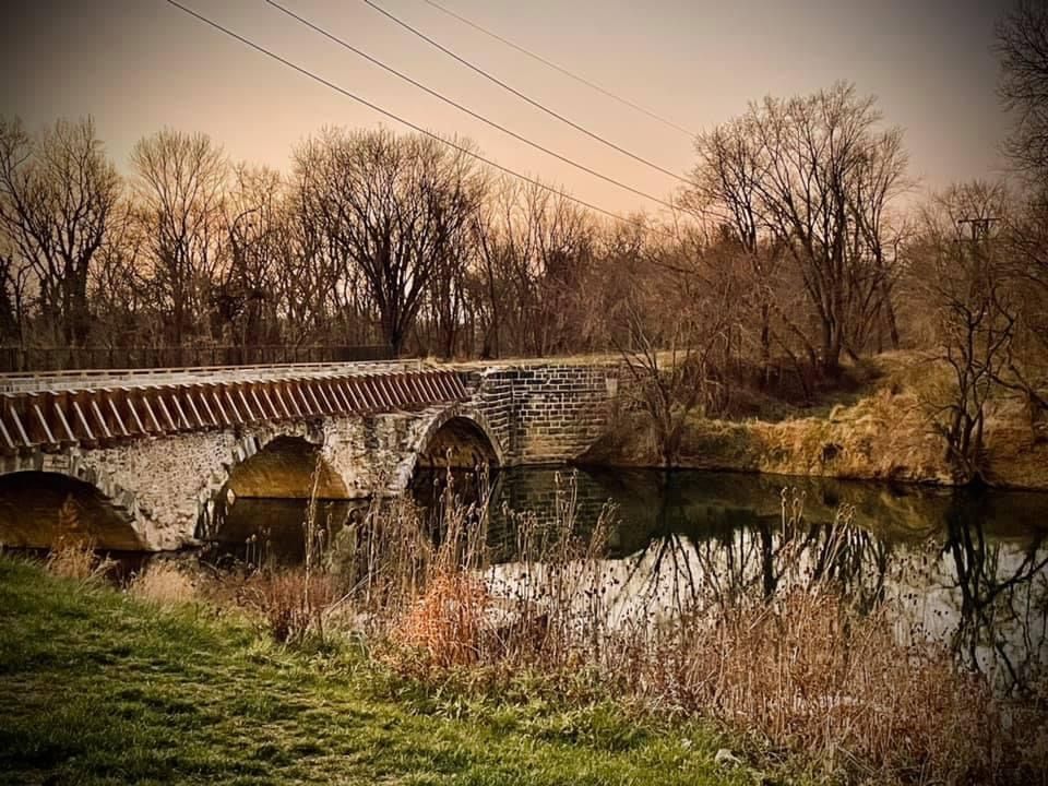 A rustic stone bridge spans across a river during autumn with bare trees lining the banks.