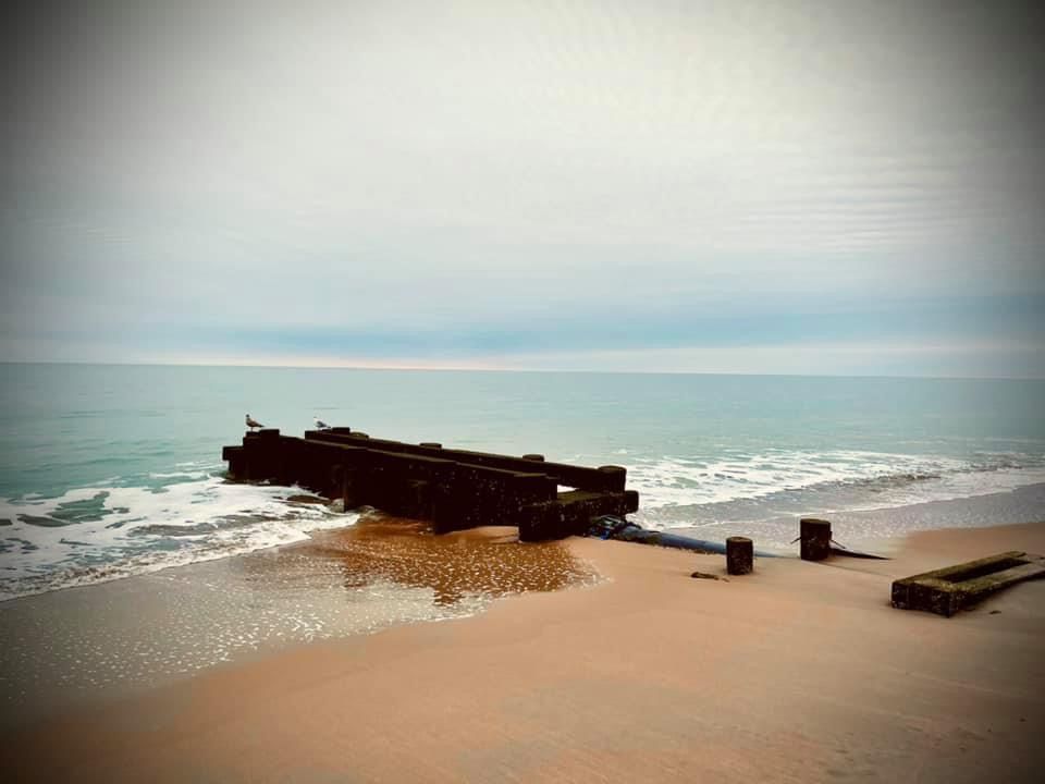 A panoramic beach scene with weathered wooden structures partially submerged in sand and surf.