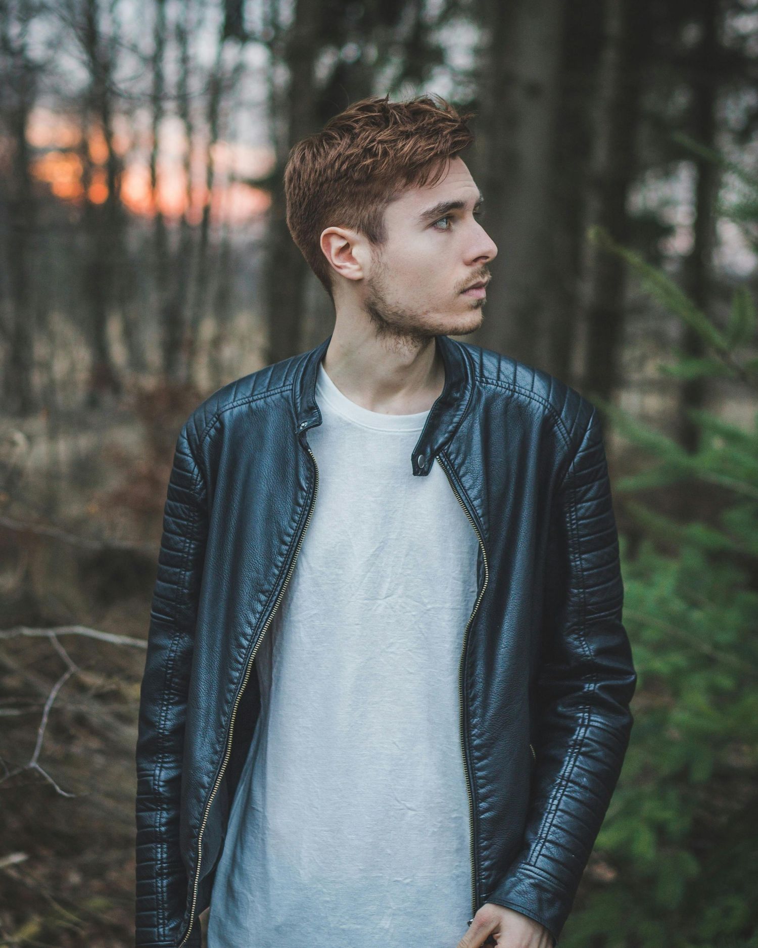 Moody portrait of person in leather jacket against forest backdrop with orange sunset.