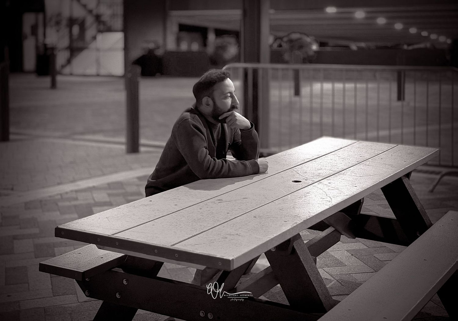 A person sits alone at a wooden picnic table in a black and white urban setting.