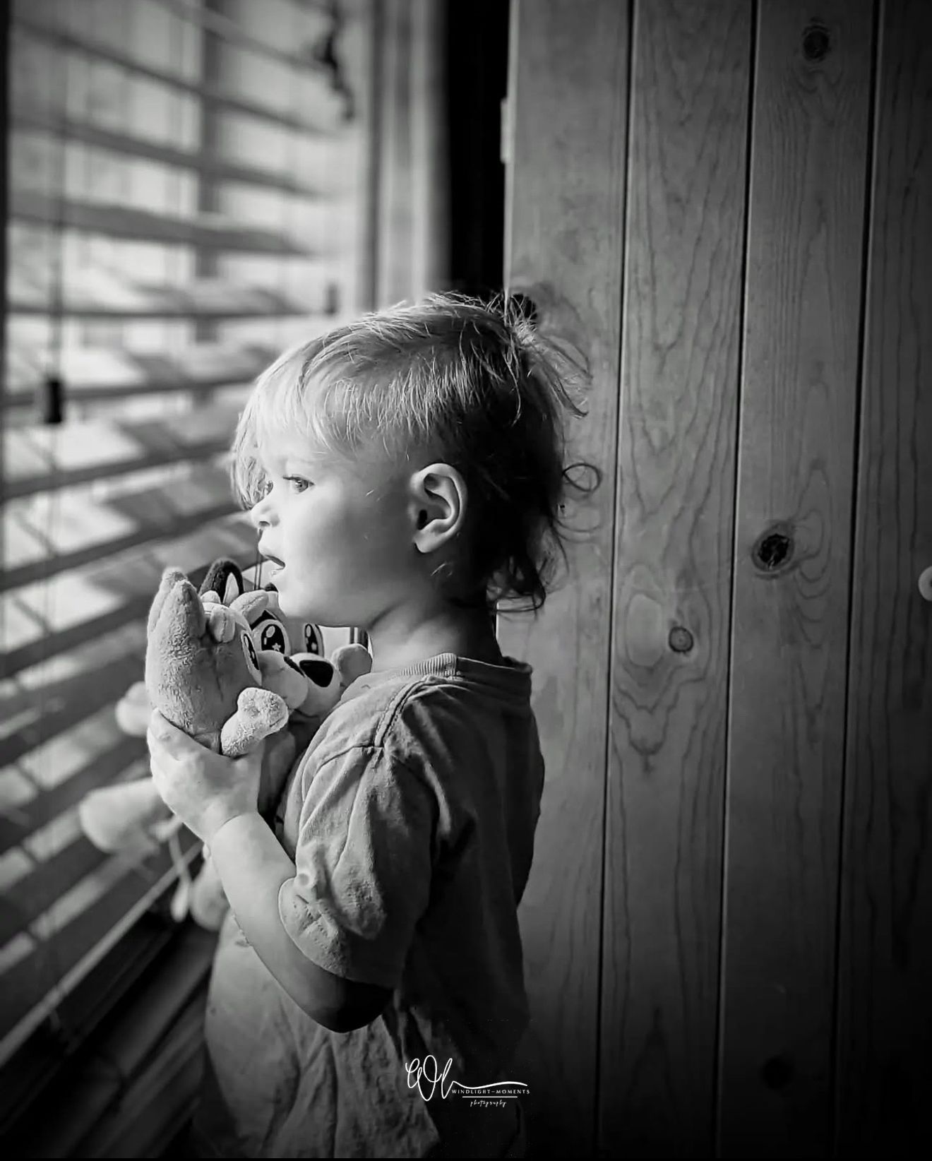 A small child in black and white peers through window blinds while holding a toy in this artistic portrait.