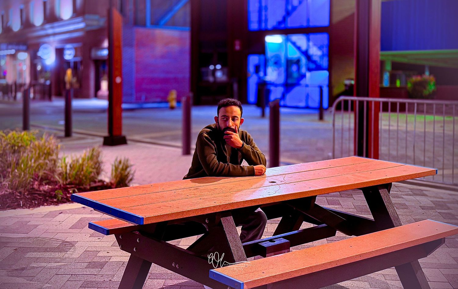 A row of wooden picnic tables illuminated by colorful blue and purple ambient lighting in an urban setting at night.