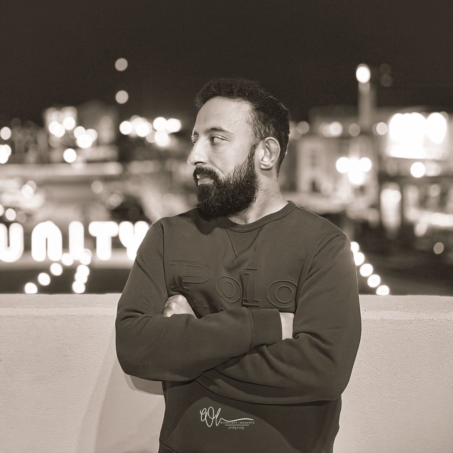 Black and white nighttime portrait of someone with a beard wearing a sweatshirt against a bokeh city lights background.