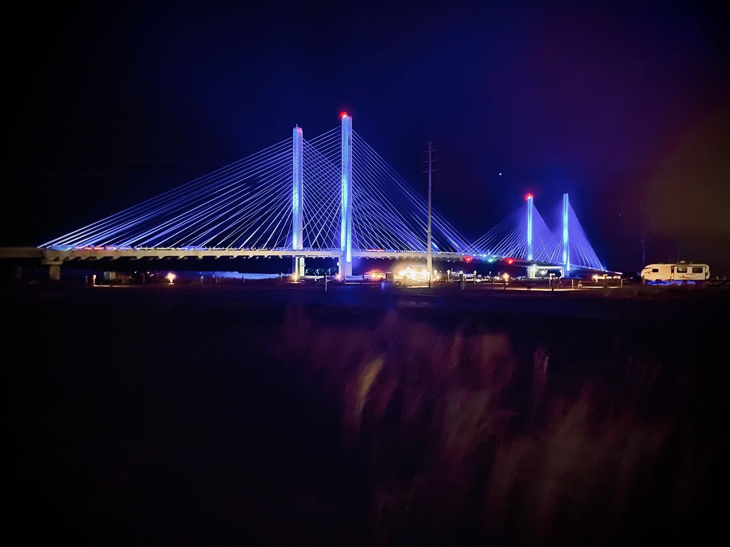 Illuminated cable-stayed bridge glowing with blue and purple lights against dark night sky.