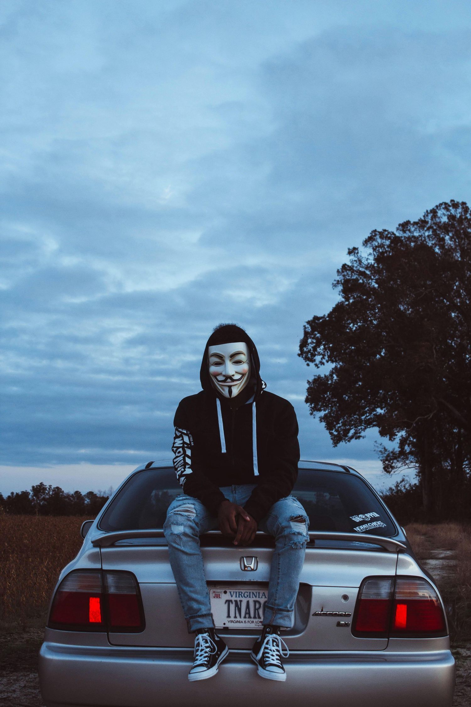 Two figures sitting on a car trunk at dusk wearing masks and hoodies.
