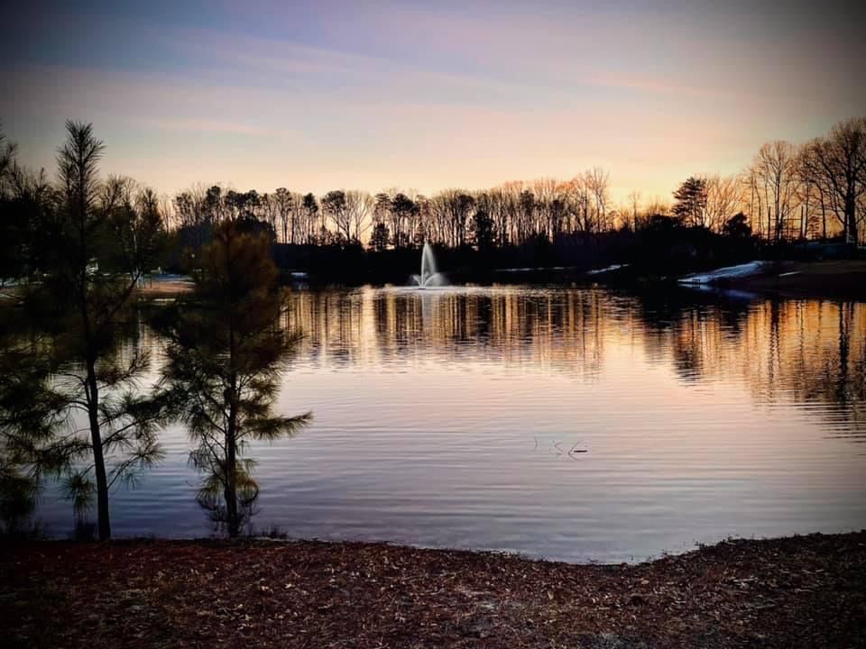 Tranquil lake at sunset with fountain and tree silhouettes along the shoreline.