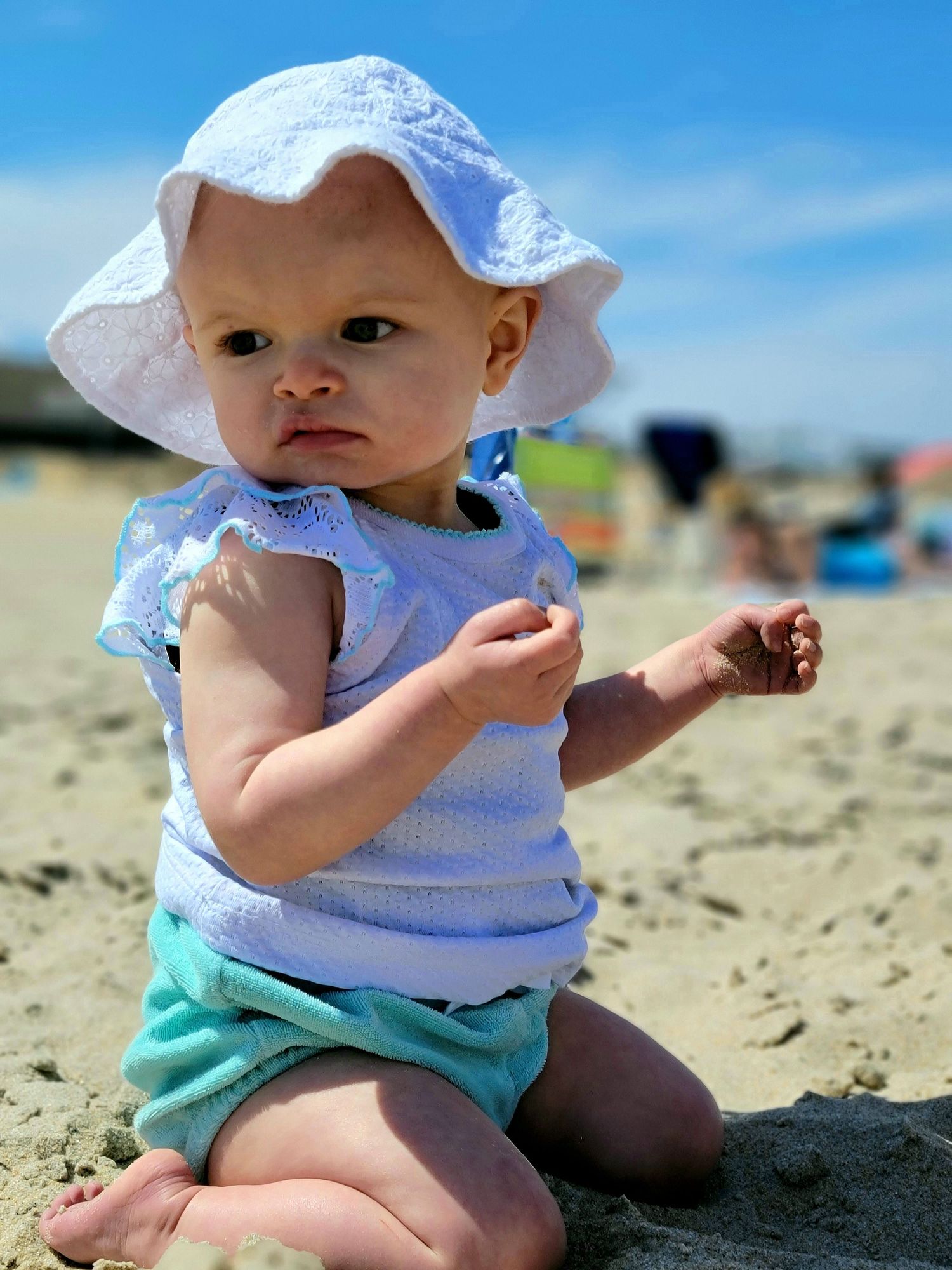 Small child playing in the sand at a sunny beach wearing a white hat.