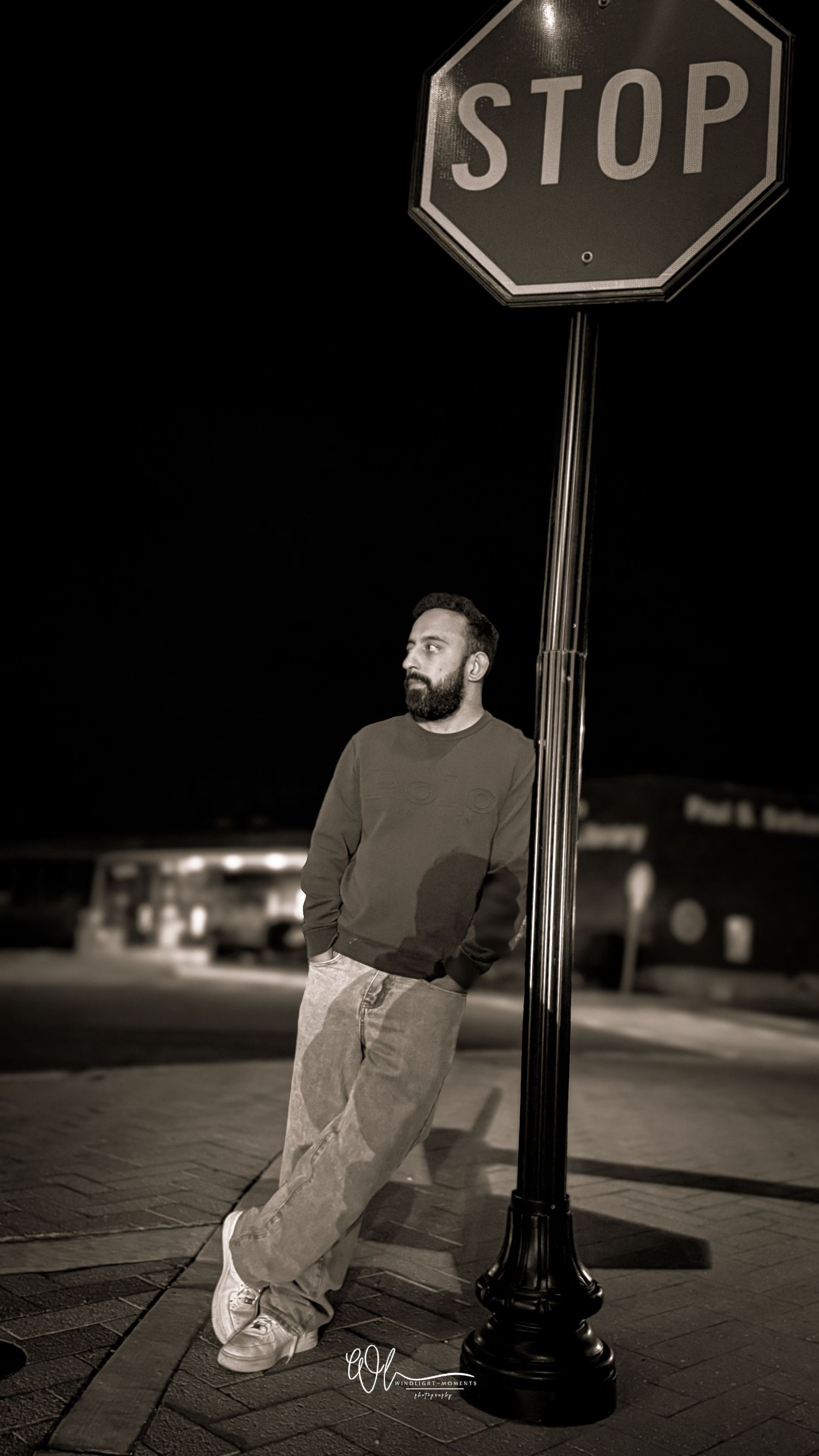 A black and white nighttime street photo of someone casually leaning against a stop sign pole on an empty sidewalk.
