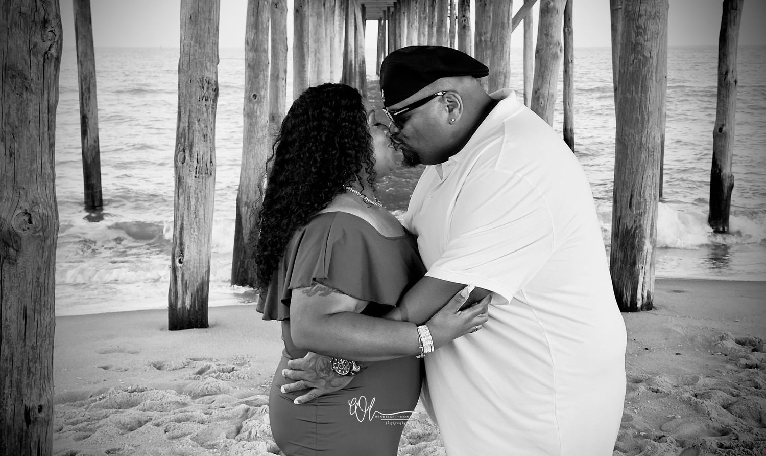 A romantic couple embraces under a wooden pier at the beach in black and white.
