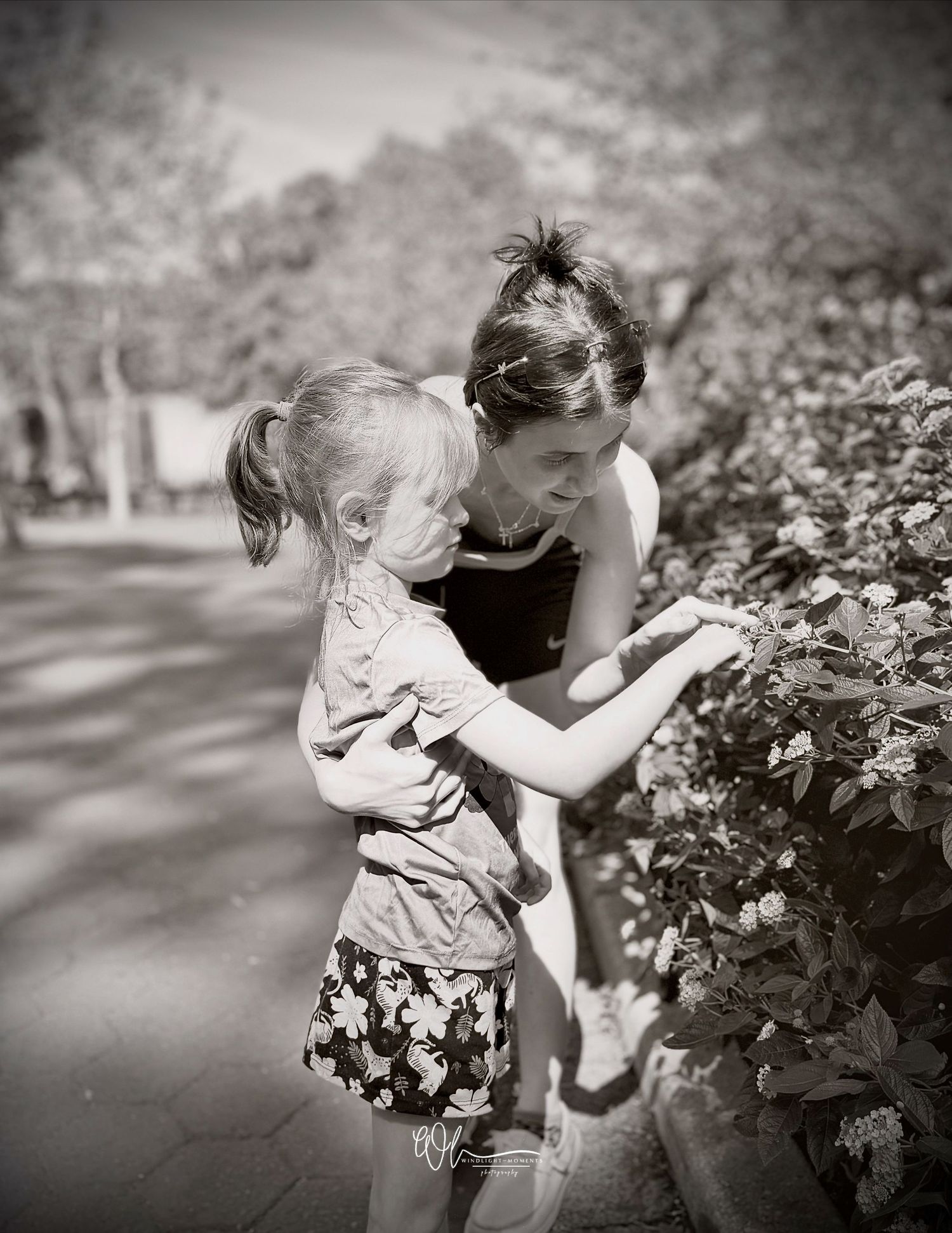 A person and child smell flowers together in a black and white garden scene.