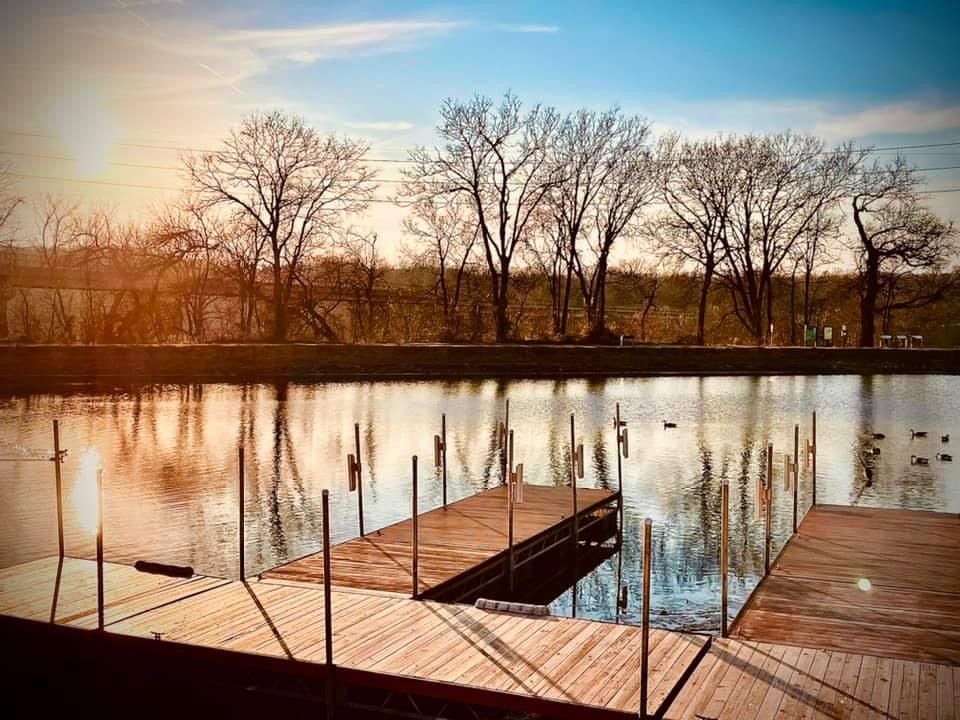 Wooden dock extends over calm water at sunset with bare trees silhouetted against the sky.