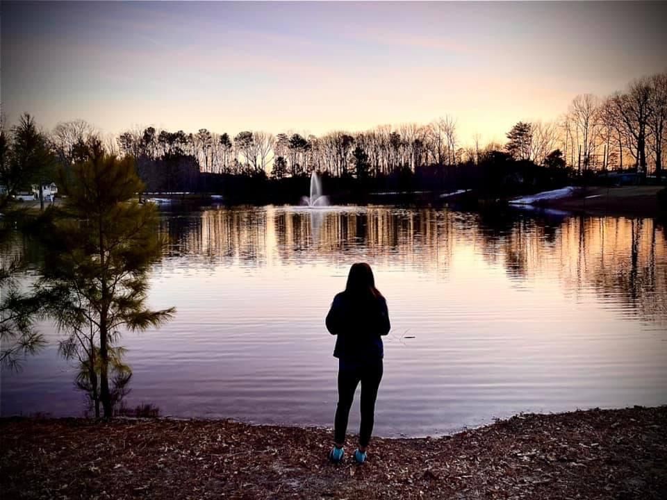 Silhouettes of people watching sunset over a calm lake with trees reflected in the water.