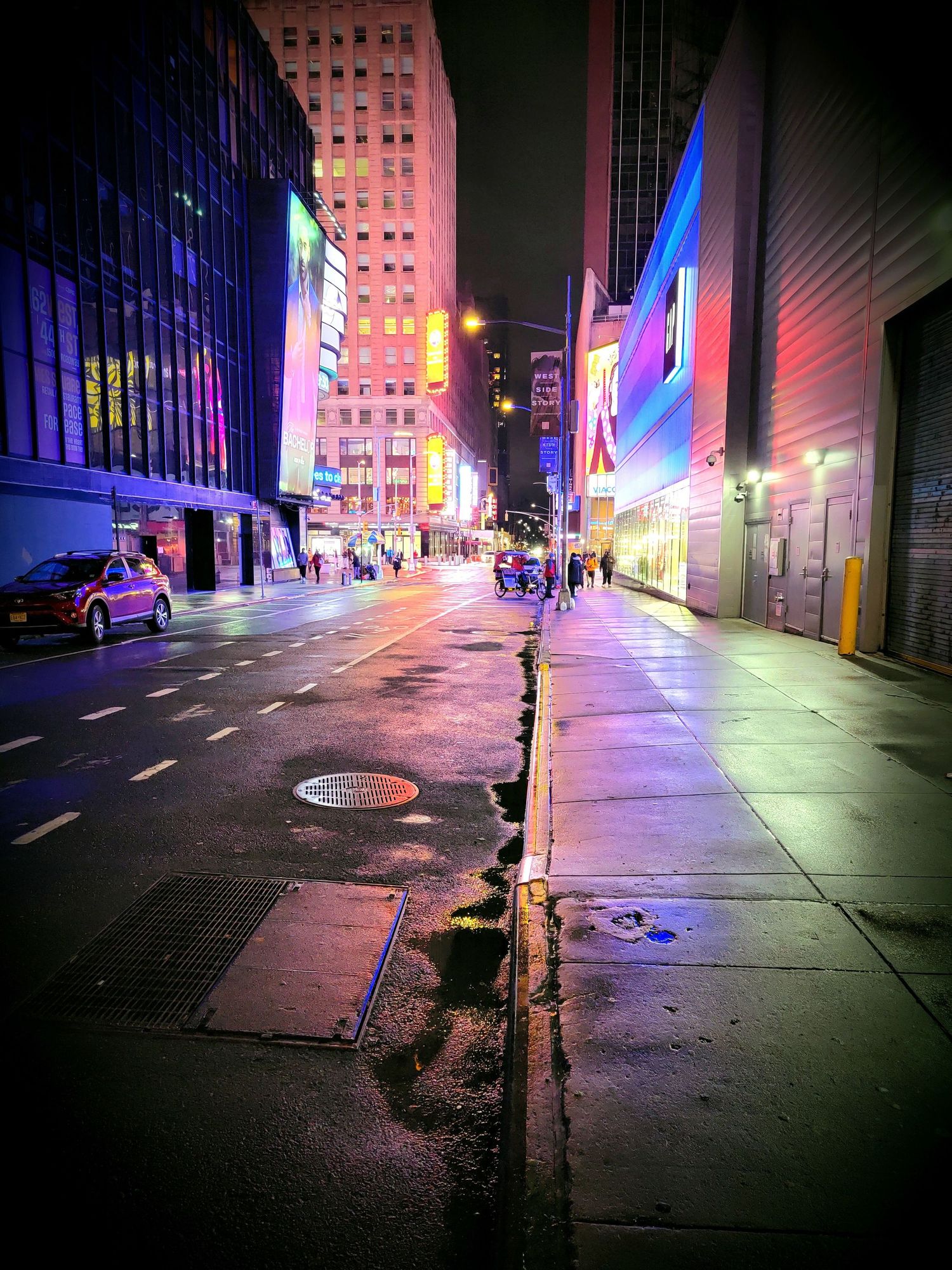 Empty city street at night with colorful neon lights casting pink and blue hues on buildings.