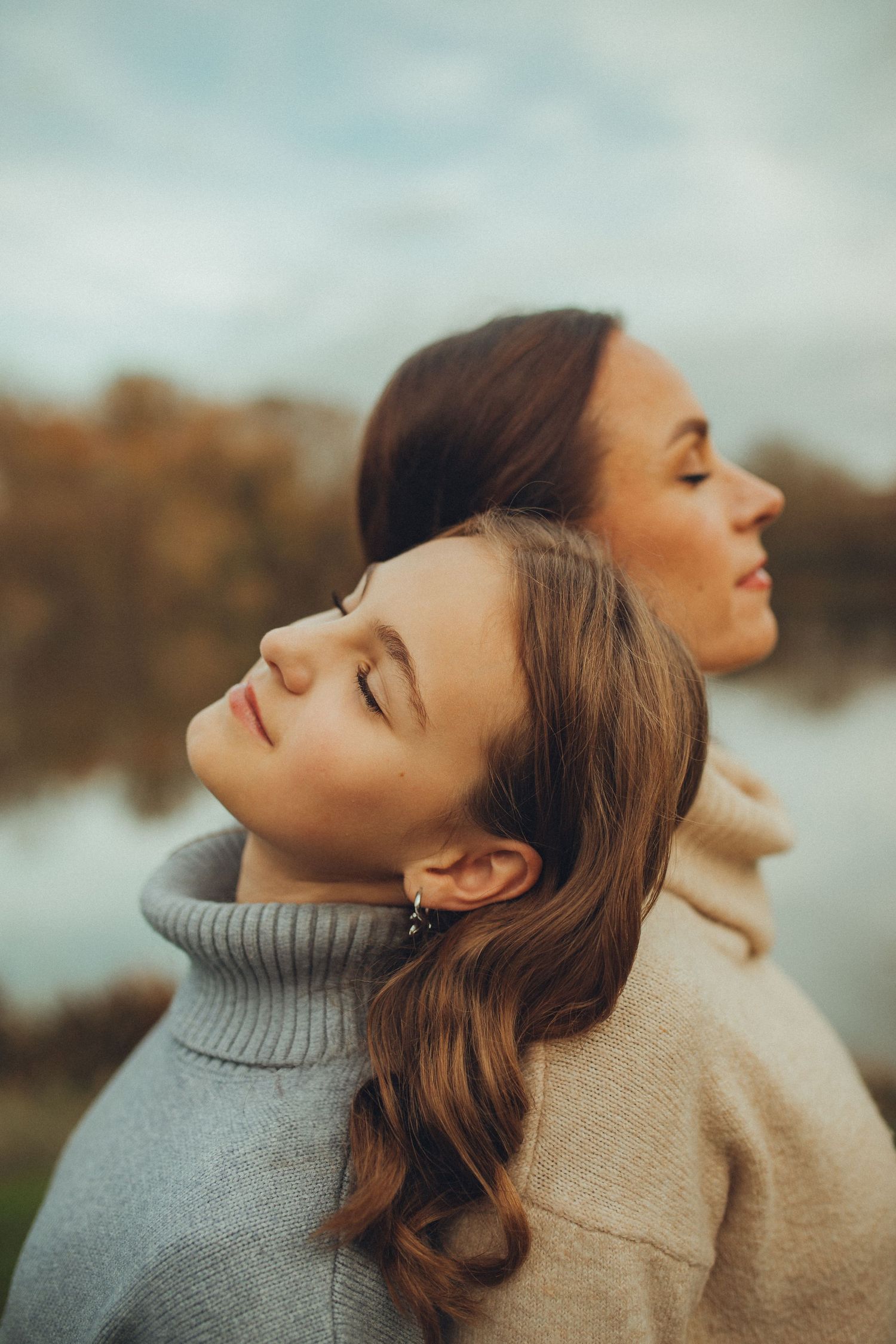 Two women in turtleneck sweaters pose with their eyes closed in a dreamy outdoor portrait.