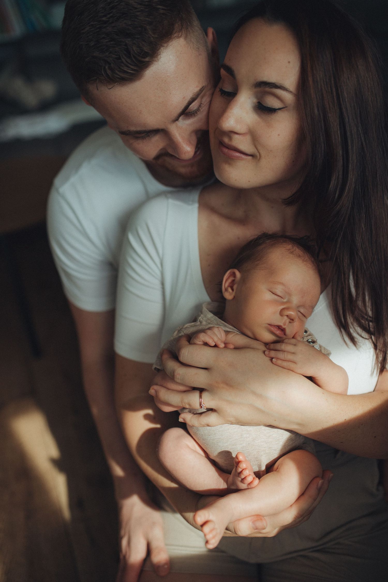 A young couple tenderly holds their sleeping newborn baby in warm natural light.