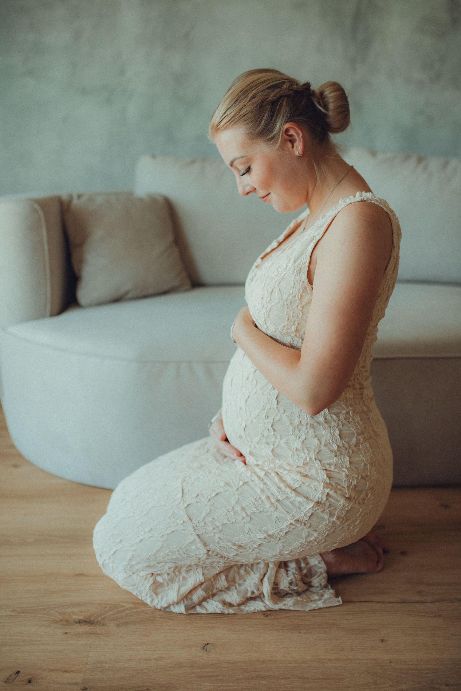 A pregnant woman in a white lace dress sits gracefully on a wooden floor in a soft, dreamy indoor setting.