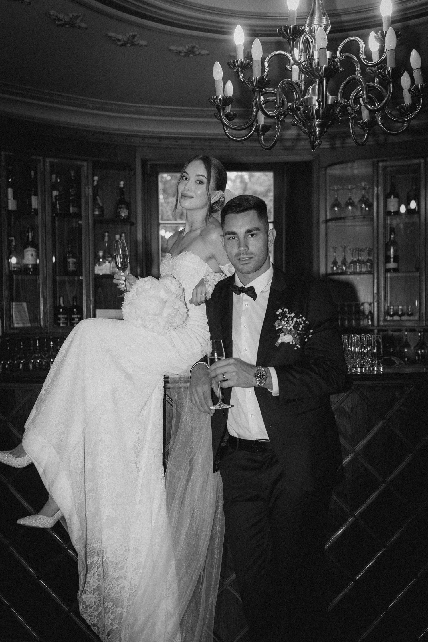Elegant couple poses in formal wedding attire near vintage bar with chandelier overhead.