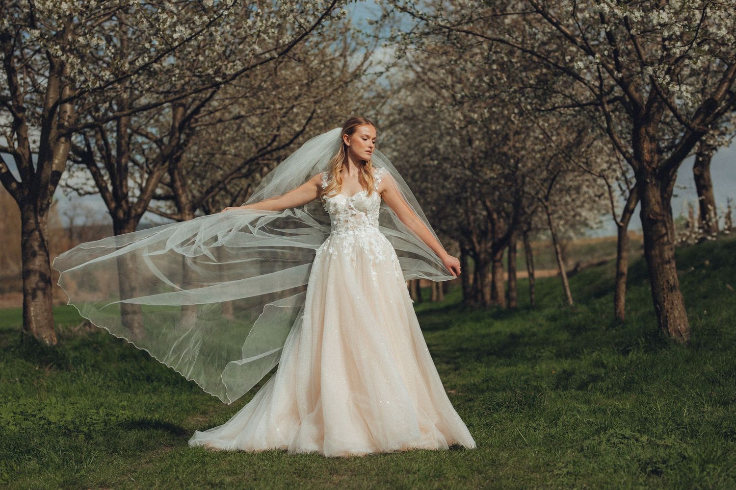 Ethereal wedding dress with delicate floral embroidery flows in a natural outdoor setting with blooming trees.