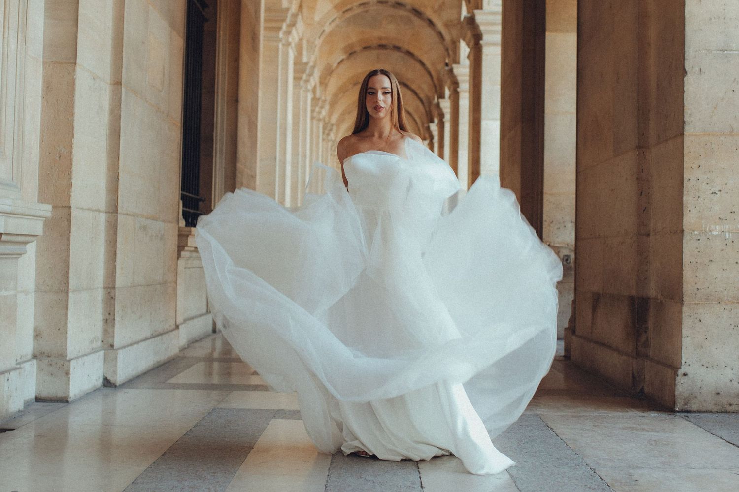 A flowing white wedding dress twirls dramatically in an elegant arched corridor with stone columns.