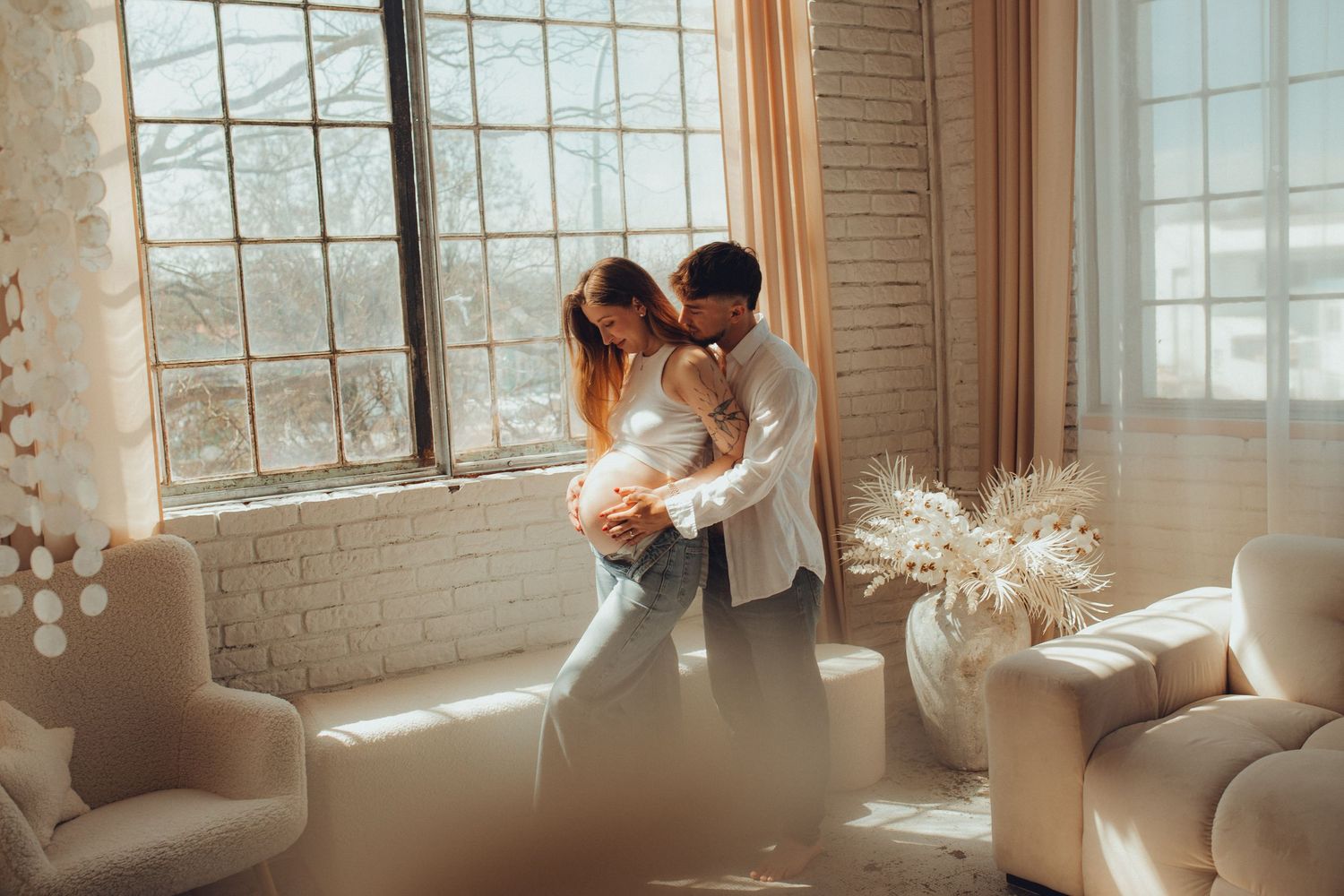 A couple in casual clothing standing together in a bright room illuminated by natural light from large windows.