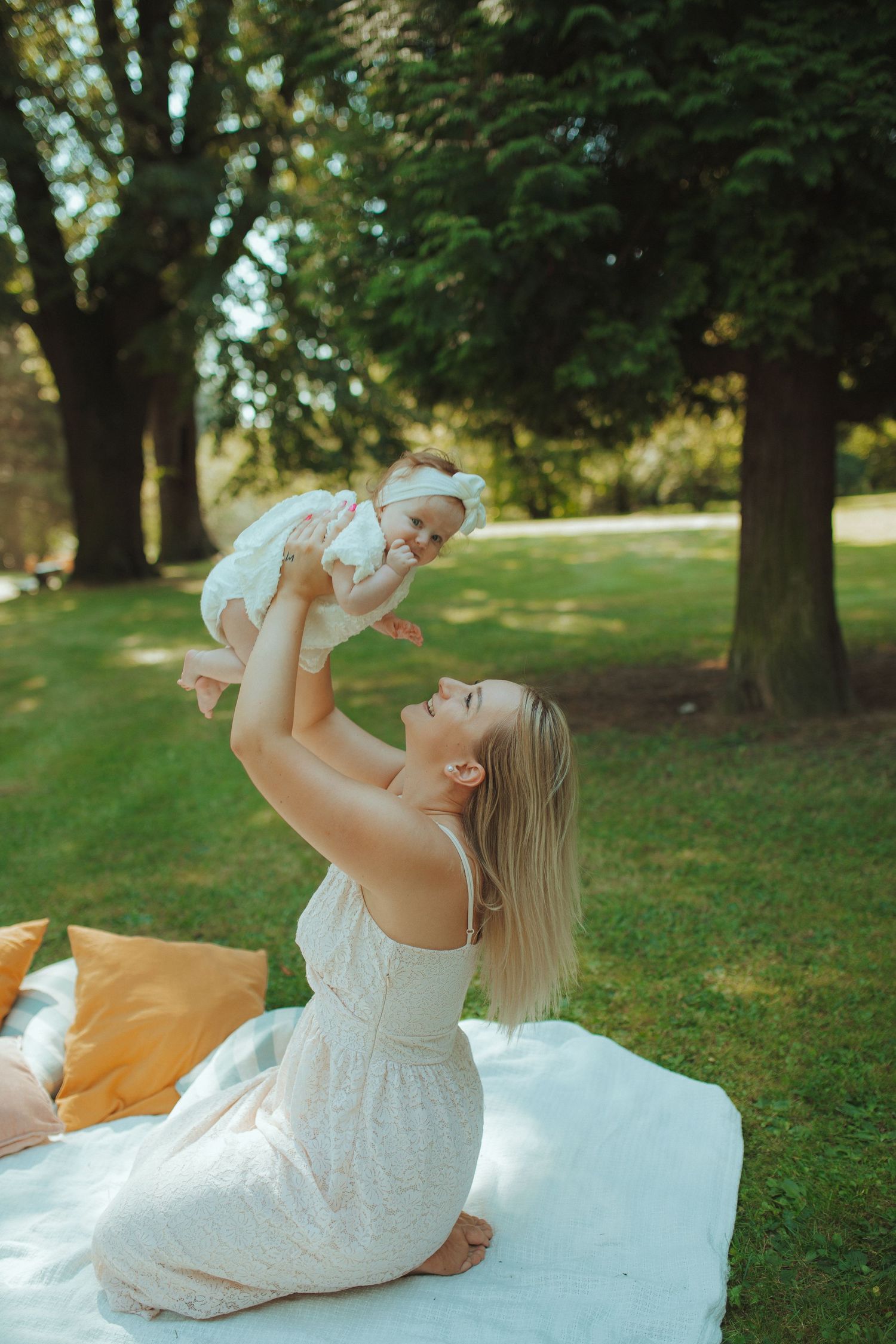 A joyful moment in a sunny park as a woman in white dress lifts a baby on a picnic blanket.