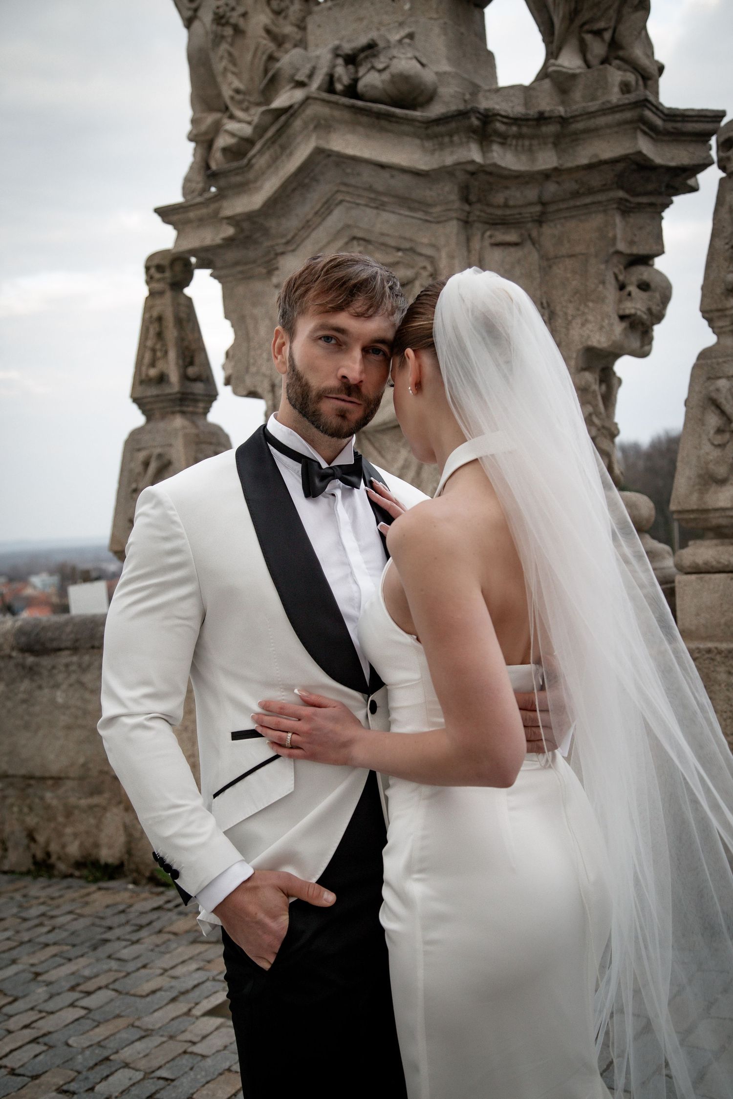 Elegant bride and groom in white tuxedo share intimate moment near ornate stone sculptures outdoors.