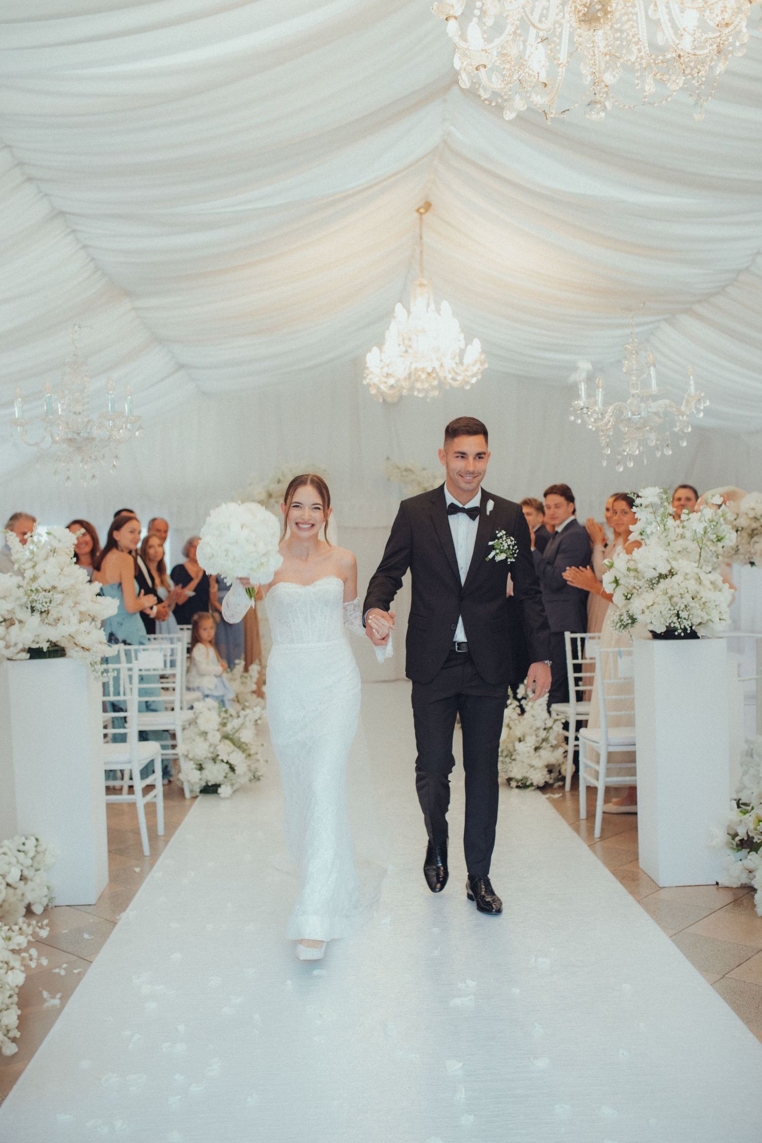 Couple walks down white aisle in luxurious tent decorated with chandeliers and floral arrangements.