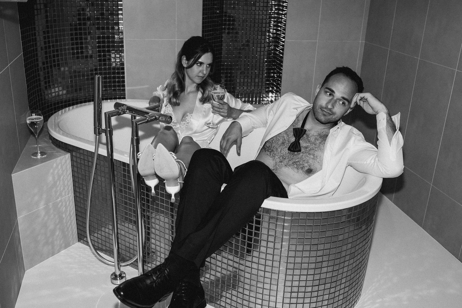 Black and white photo of couple relaxing in modern bathtub wearing formal attire.
