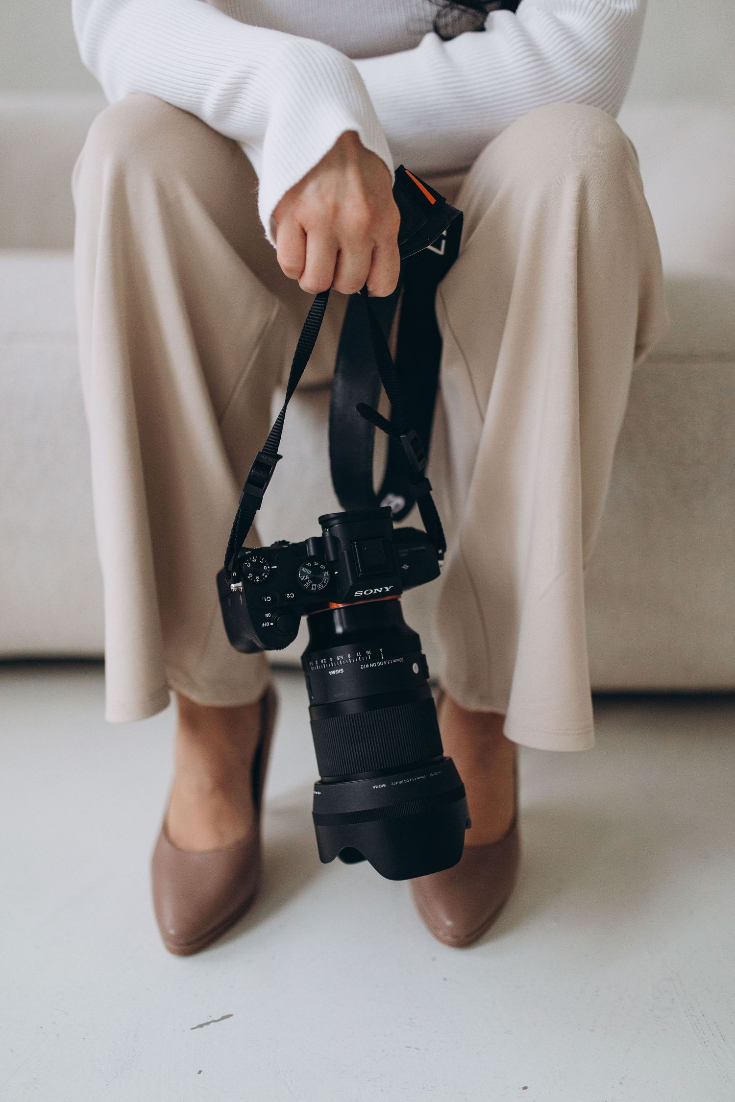 Close-up of hands holding professional camera while sitting in beige pants and brown shoes.
