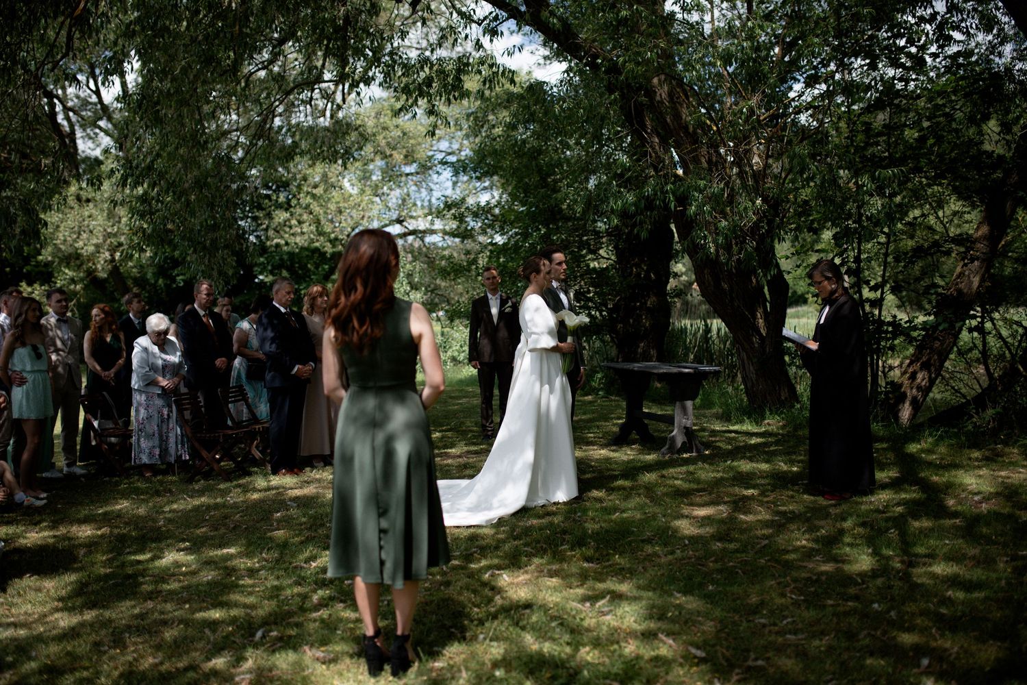 Outdoor wedding ceremony in a lush green forest setting with guests gathered around the bride and groom.