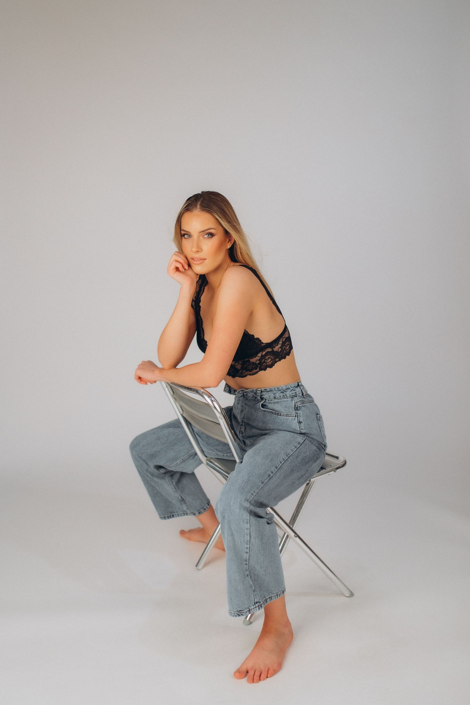 Model in casual denim and black top poses on chair in studio setting.