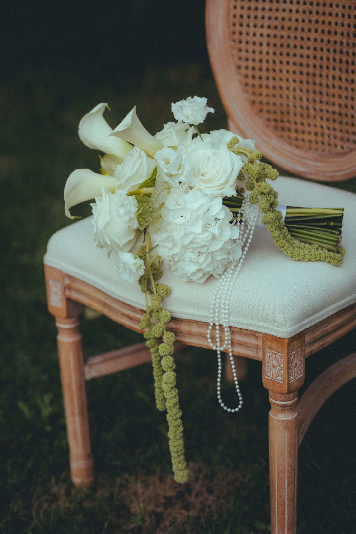 Elegant bridal bouquet of white roses and calla lilies with pearl necklace on a vintage wooden chair.