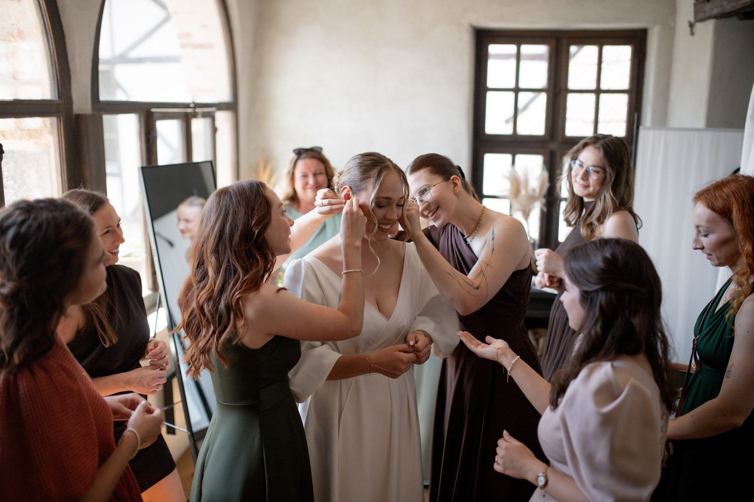Bride surrounded by bridesmaids in elegant dresses, sharing a joyful moment while getting ready for the wedding.
