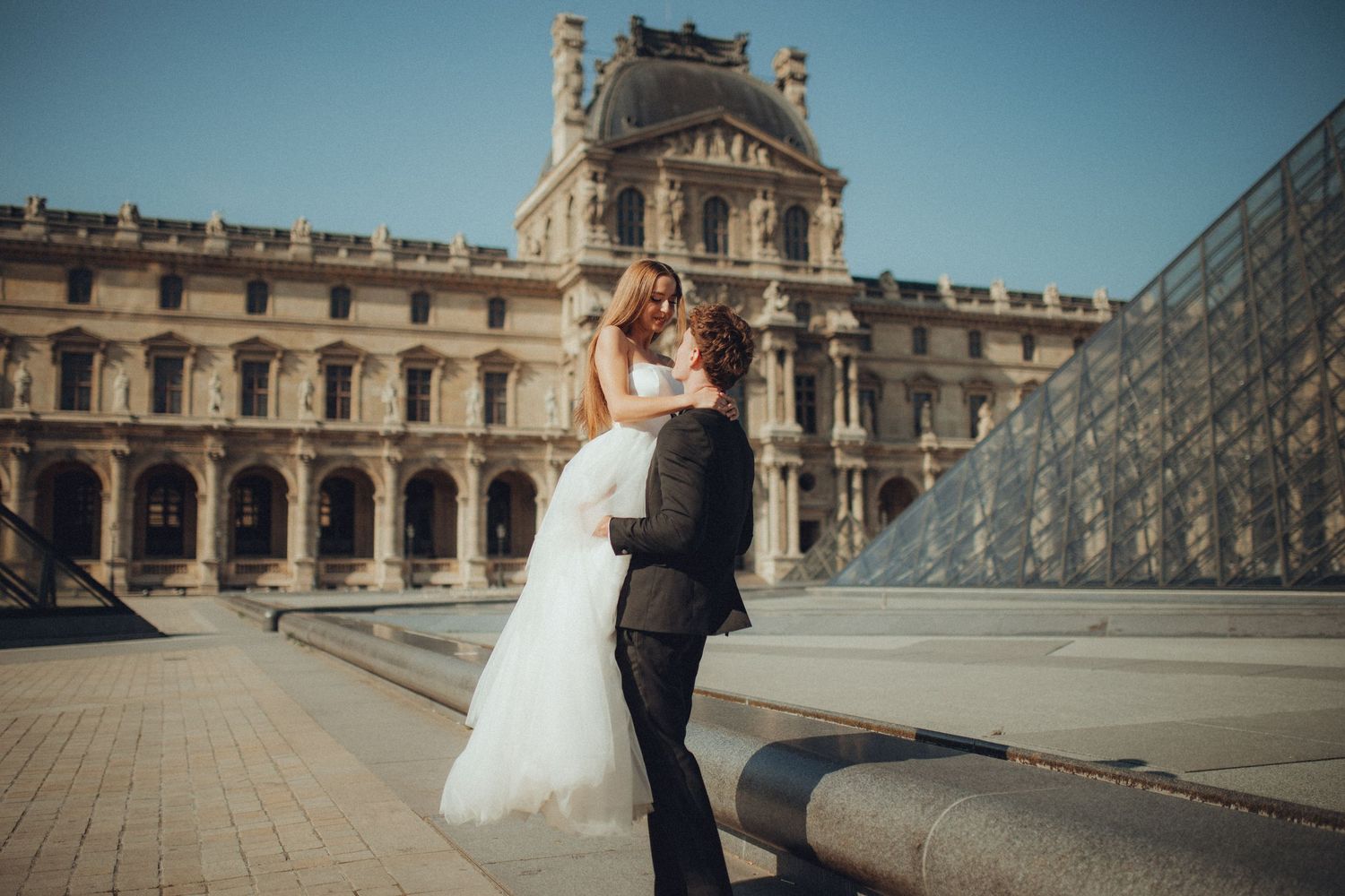A romantic wedding couple embraces in front of the iconic Louvre Museum pyramid in Paris on a sunny day.