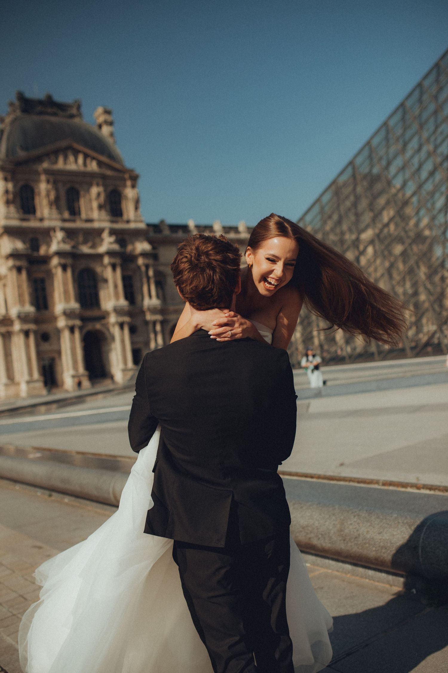 Wedding couple embraces outside the Louvre Museum in Paris with iconic glass pyramid in background.