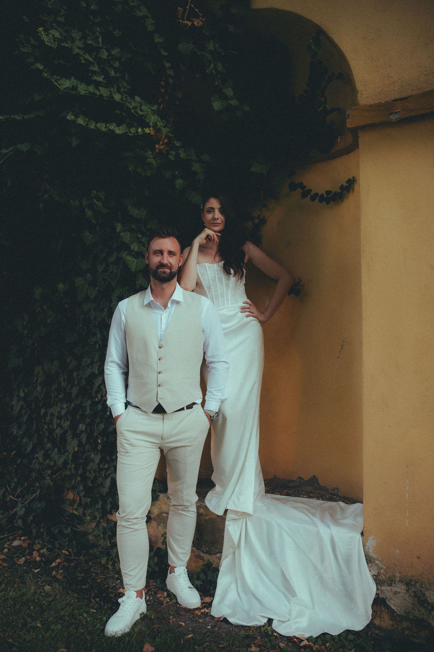 Elegant bride and groom posing outdoors near ivy-covered wall and yellow arch in matching white wedding attire.
