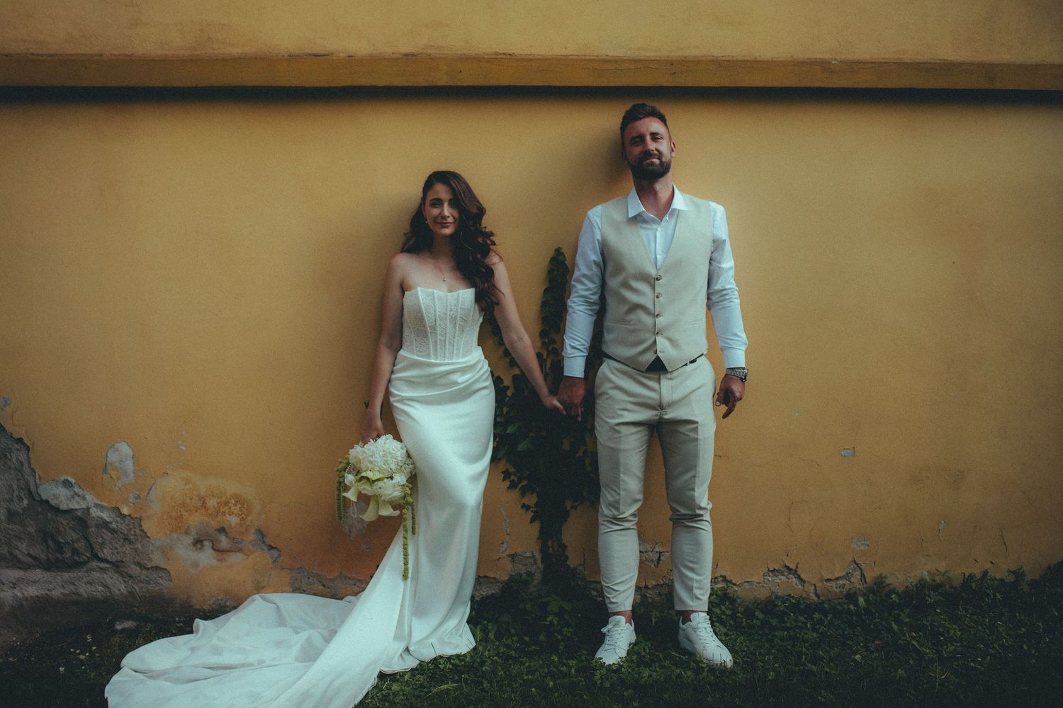 Bride in white gown and groom in beige suit hold hands against a yellow wall outdoors.