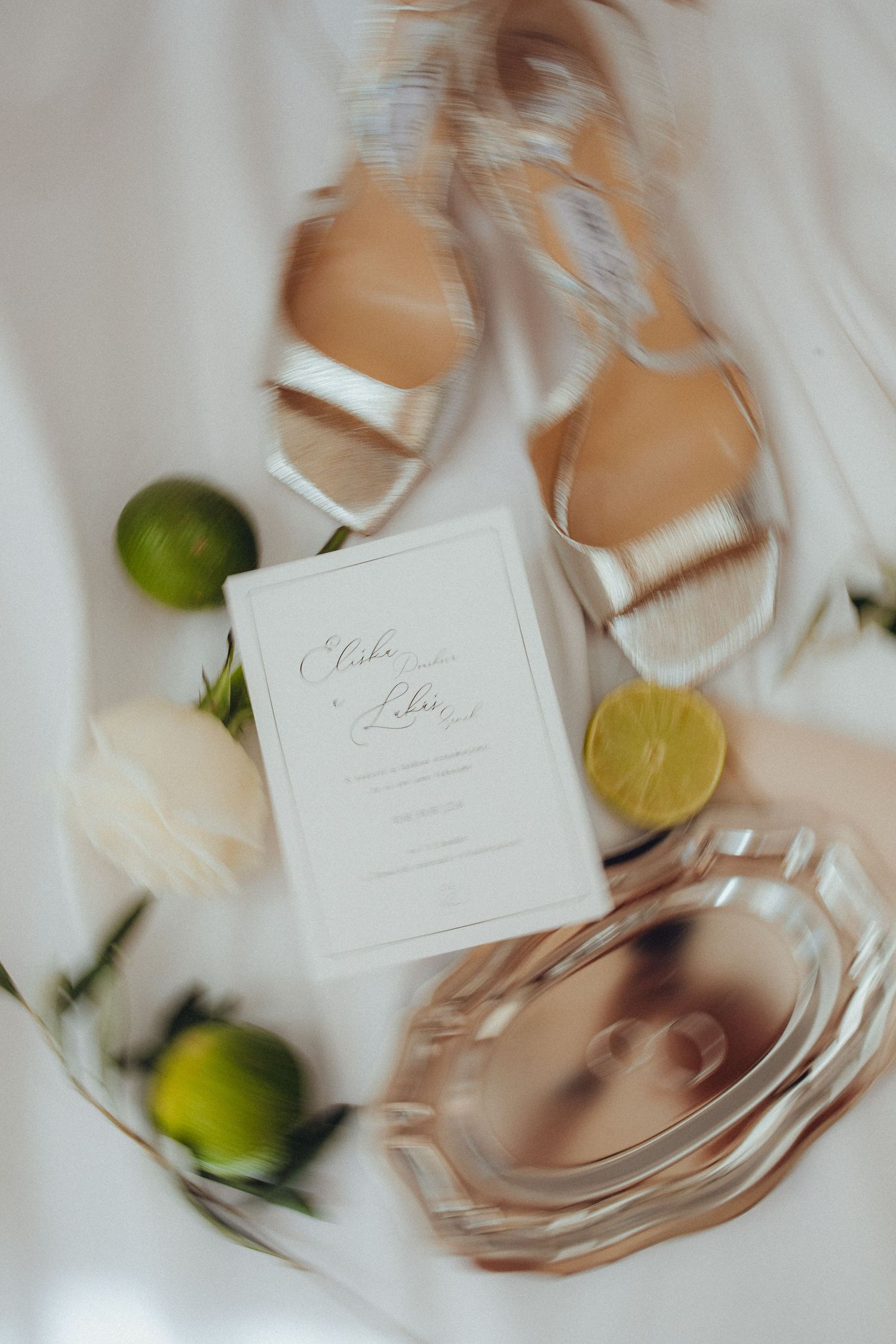 Elegant wedding flat lay with gold heels, white invitation, limes, and roses on a white fabric backdrop.