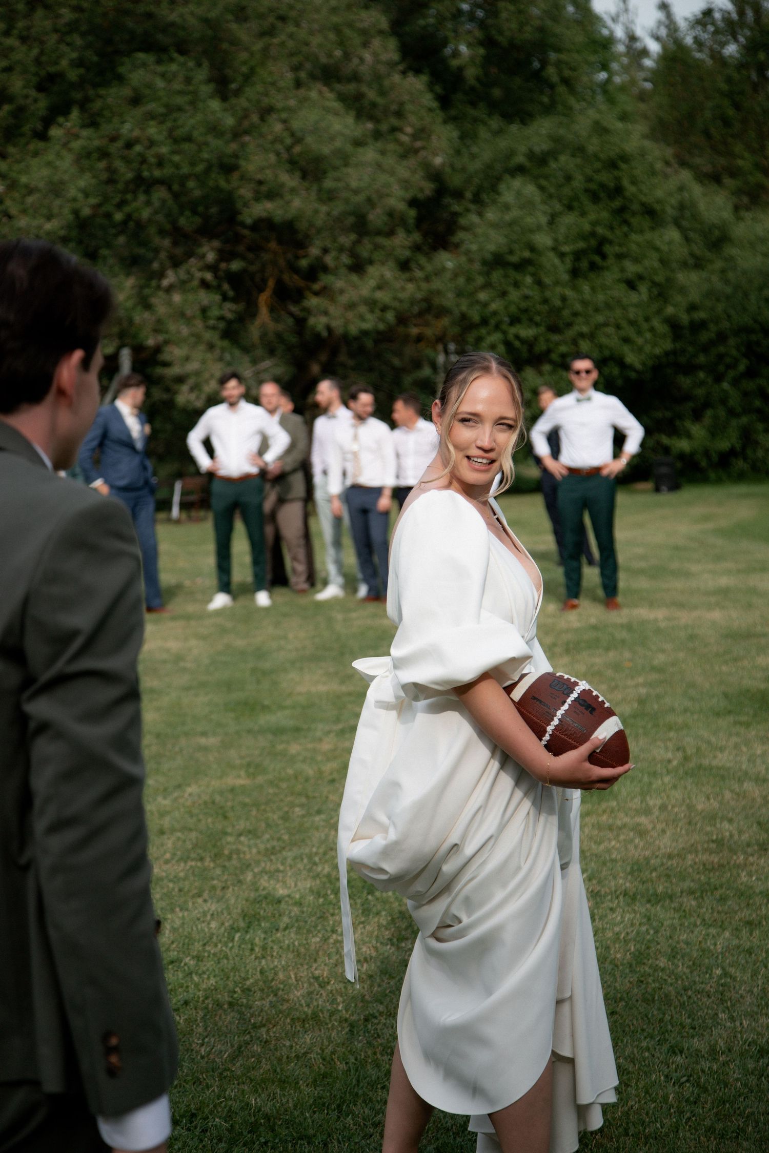 Bride in white dress holding football with groomsmen standing in background on green lawn.