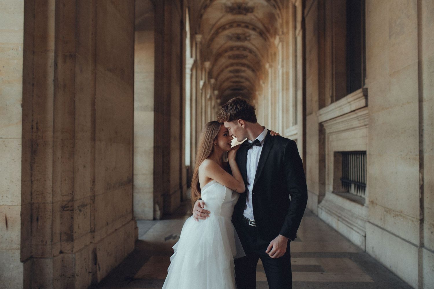 Bride and groom share a romantic moment in an elegant stone hallway with arched columns.