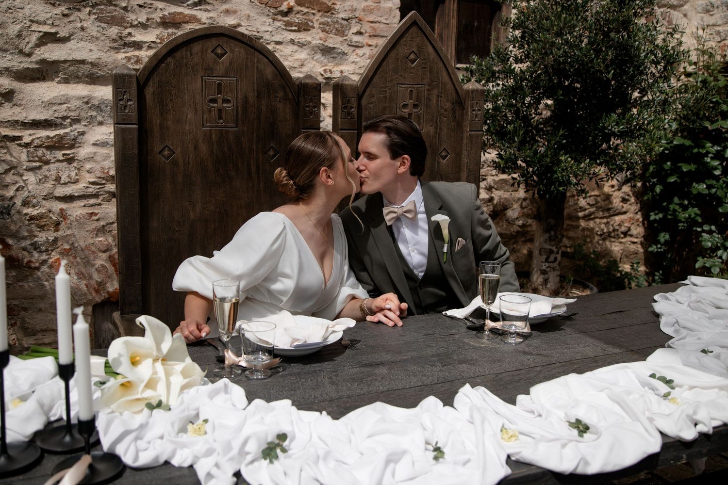 Bride and groom share a kiss at a rustic stone wedding venue table adorned with white flowers and candles.