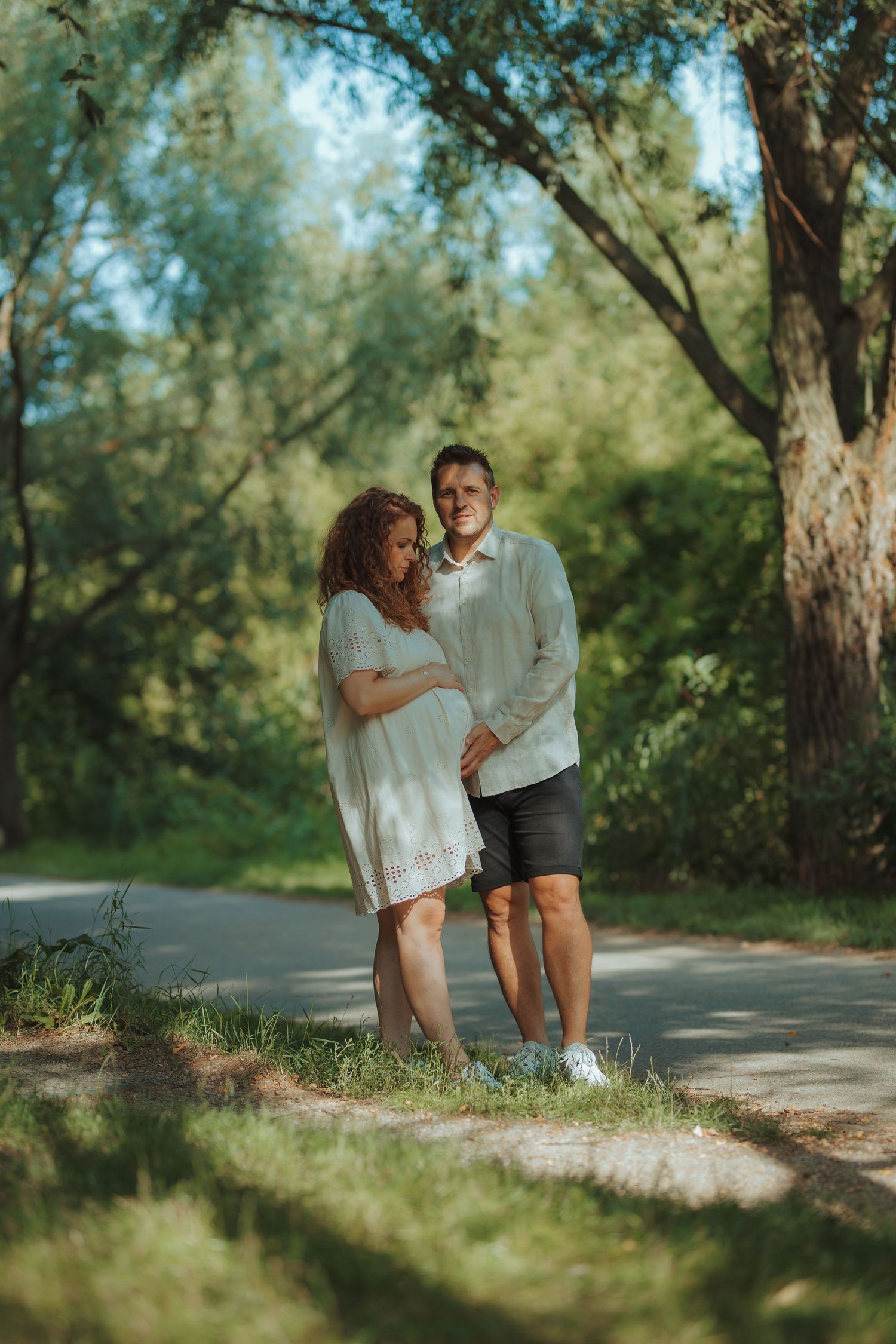 Couple in white and dark clothing sharing intimate moments along a tree-lined path outdoors.