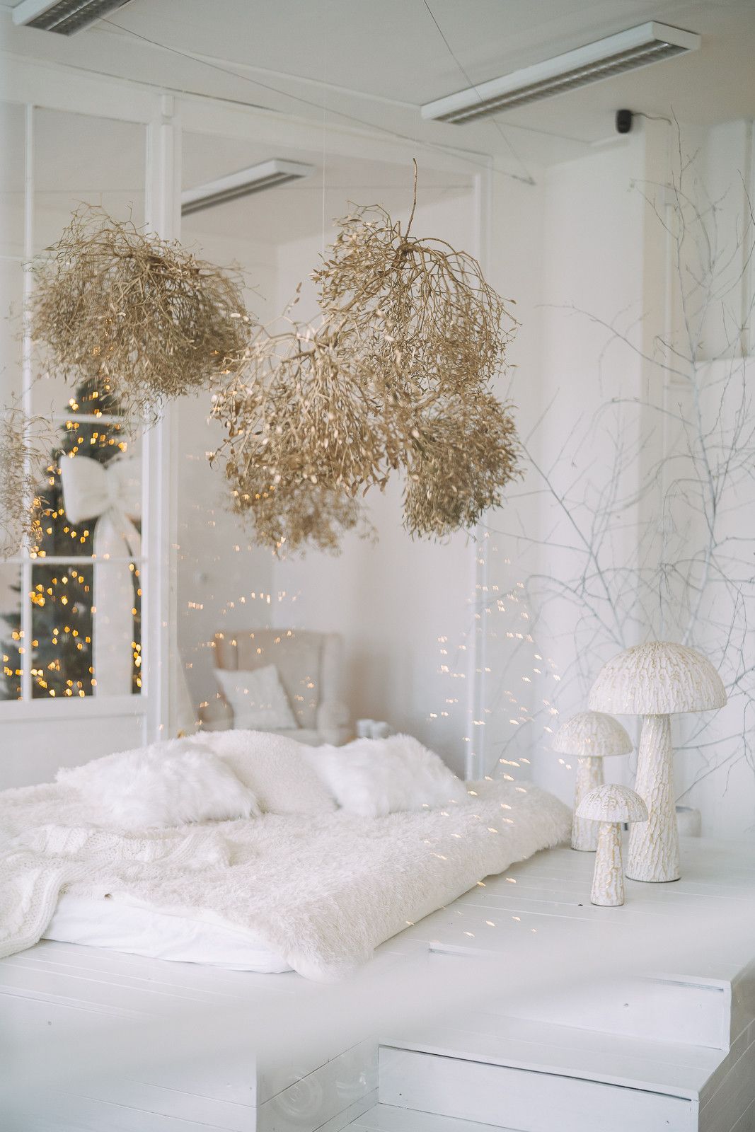 A serene all-white bedroom with dried floral arrangements hanging from the ceiling and twinkling string lights creating ambiance.