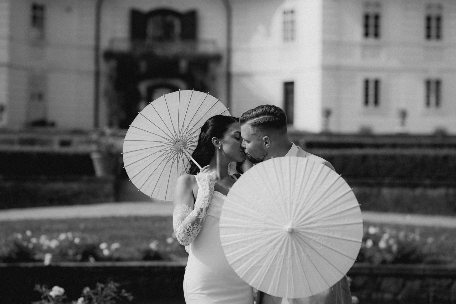 Black and white photo of a couple kissing behind white parasols in an elegant garden setting.