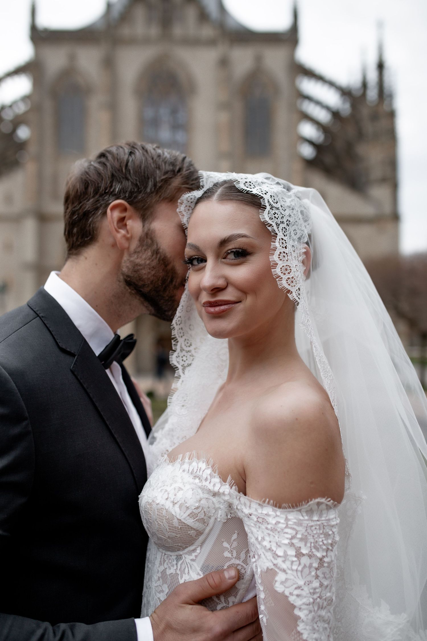 A bride in lace veil and gown smiles as groom in black tuxedo kisses her cheek before a Gothic cathedral.